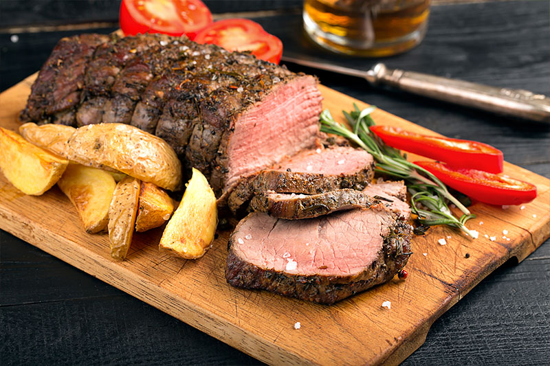 Corte de carne para churrasco alcatra assada, fatiada, posicionado em cima de tábua de madeira com batatas, pimentas e ervas ao lado da carne. 