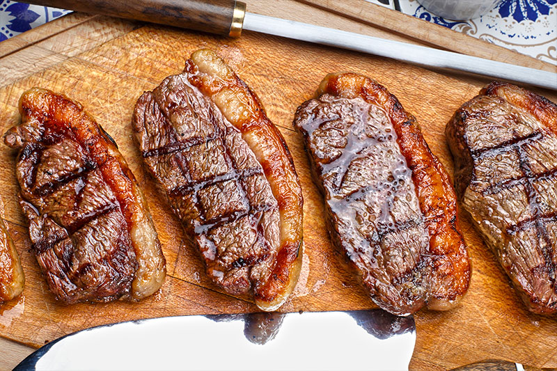 Carne para churrasco - quatro bifes de picanha bovina, suculentos, assados, posicionados em cima de tábua de madeira. 